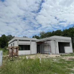 Niedokończony dom z białych bloczków, widoczne rusztowanie, palety i bujna roślinność w otoczeniu. Etap budowy w słoneczny dzień.