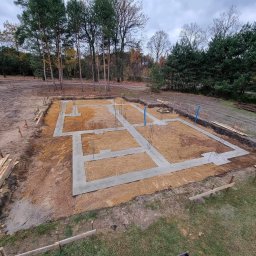 Usługi Ogólnobudowlane Błażej Kański - Zalane betonem fundamenty domu jednorodzinnego z wystającymi prętami zbrojeniowymi. Widok z góry na wykopane i przygotowane podłoże z deskami szalunkowymi.