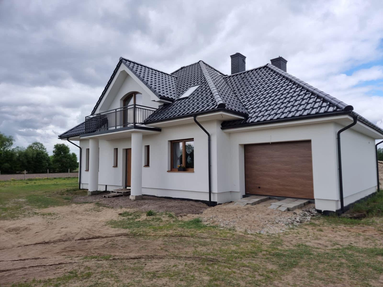 Nowoczesny dom jednorodzinny z balkonem i garażem, wykończony w bieli z ciemną dachówką. Widok z boku, na pierwszym planie teren przygotowany pod trawnik.