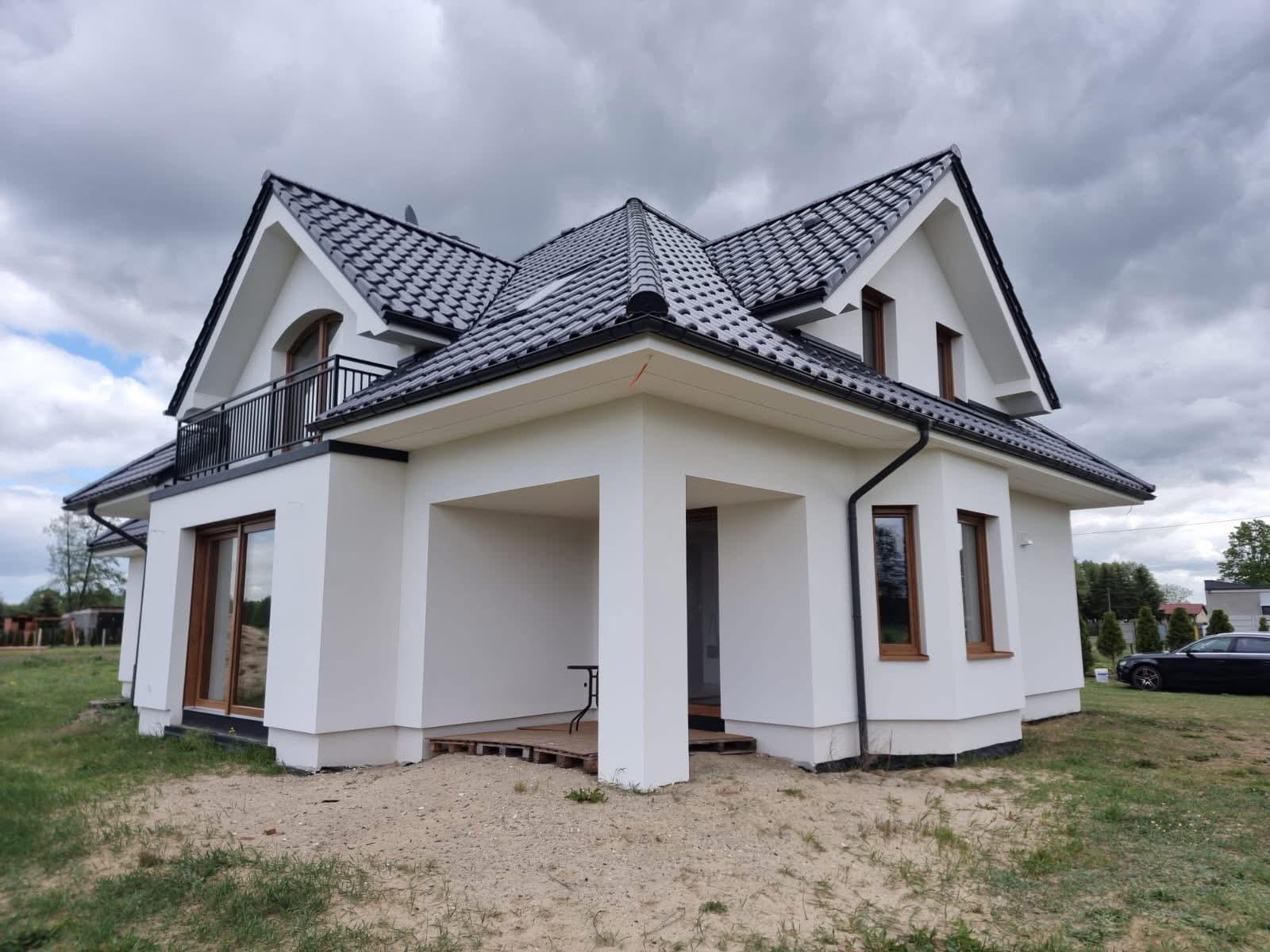 Biały, nowoczesny dom z czarnym dachem i balkonem, widok z zewnątrz w pochmurny dzień. Widoczne elementy wykończeniowe i otoczenie budynku.