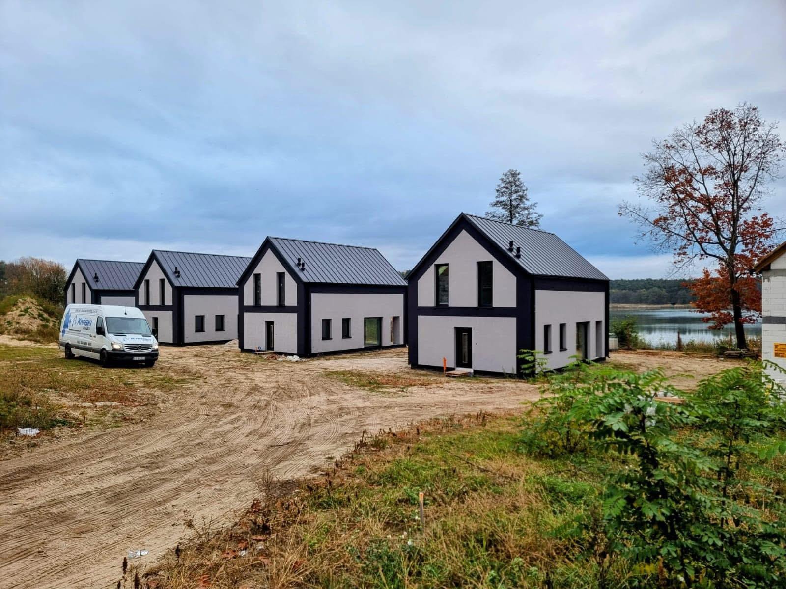 Rząd identycznych, nowoczesnych domów z szarą blachodachówką i czarnymi obramowaniami okien, z firmowym vanem z boku, na tle jeziora i pochmurnego nieba.