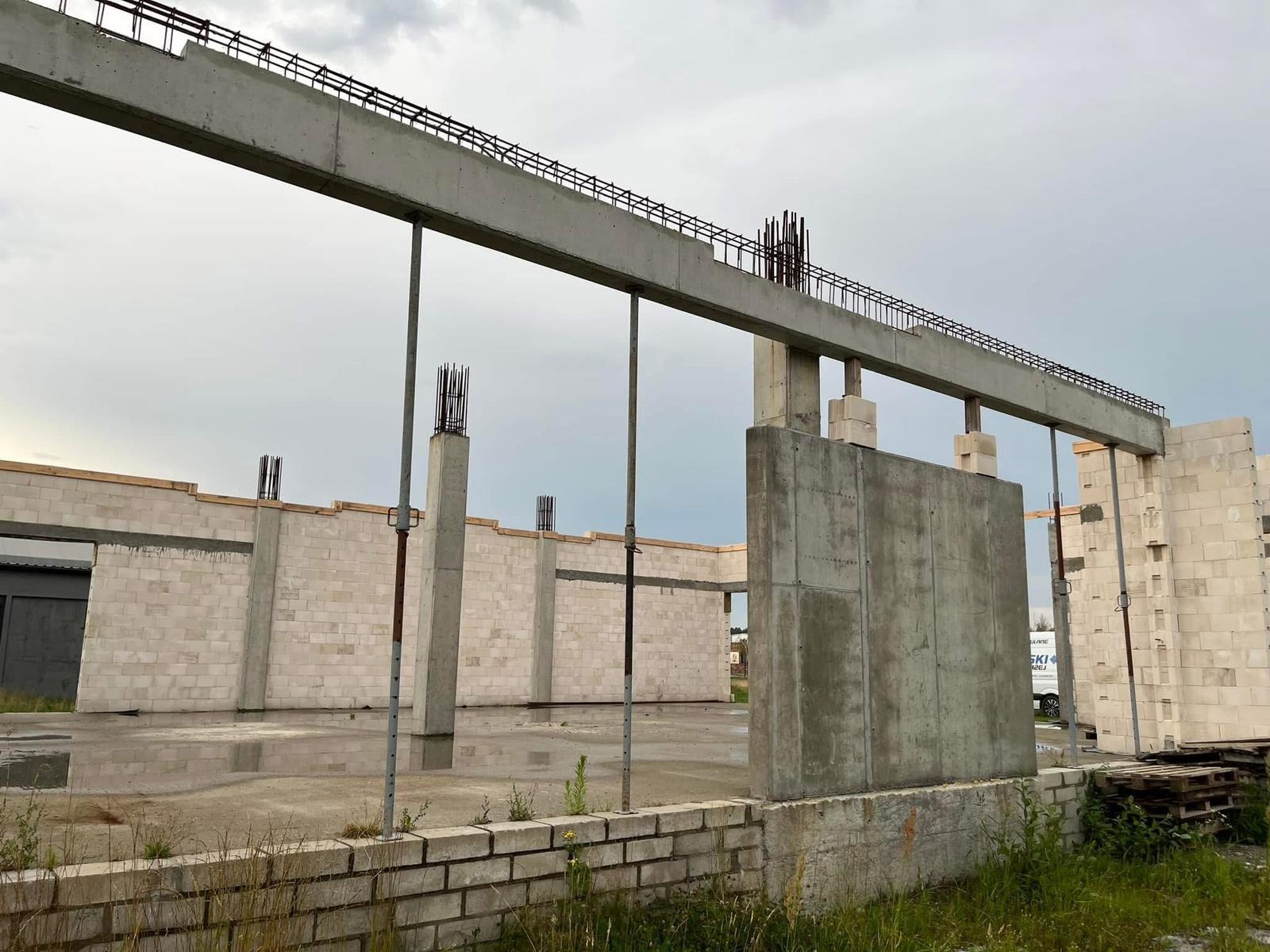 Budynek w stanie surowym otwartym: ściany z betonu komórkowego, żelbetowe słupy ze zbrojeniami, betonowe nadproża ze stalą zbrojeniową. Widoczne podpory i fundamenty.