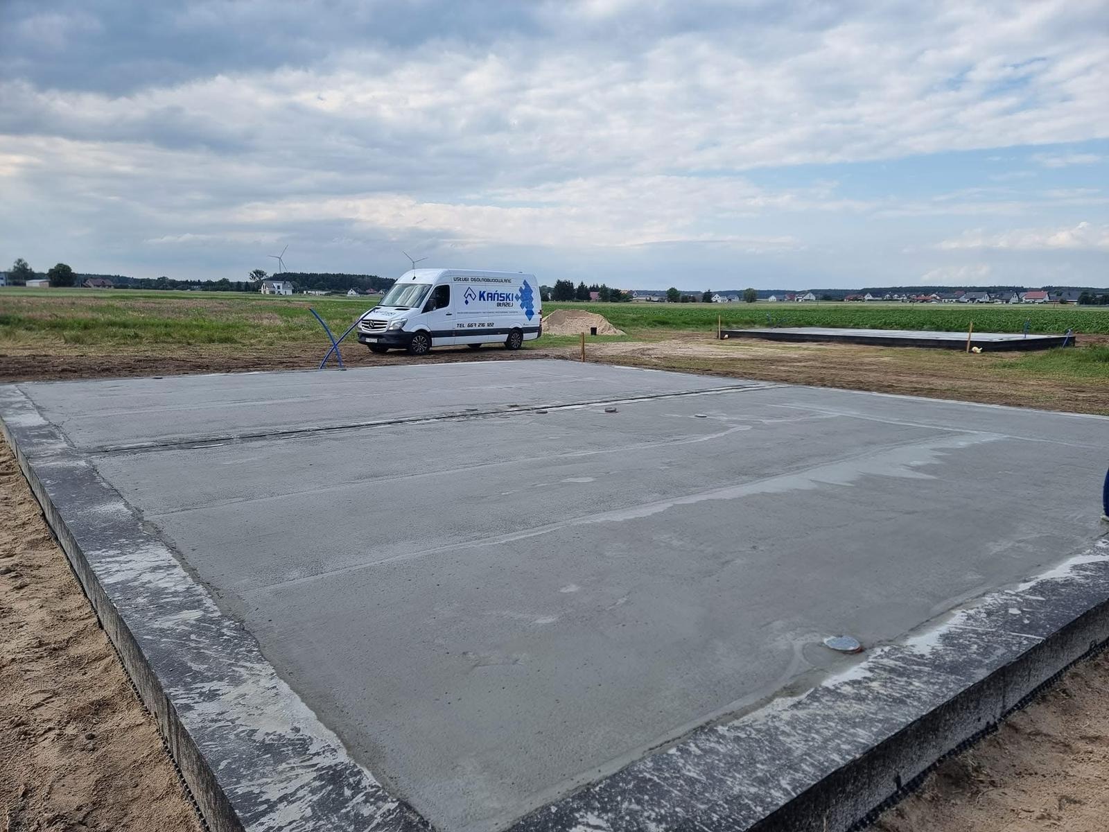 Świeżo wylany fundament pod budynek, z widocznym firmowym vanem w tle na tle krajobrazu polnej działki w słoneczny dzień.