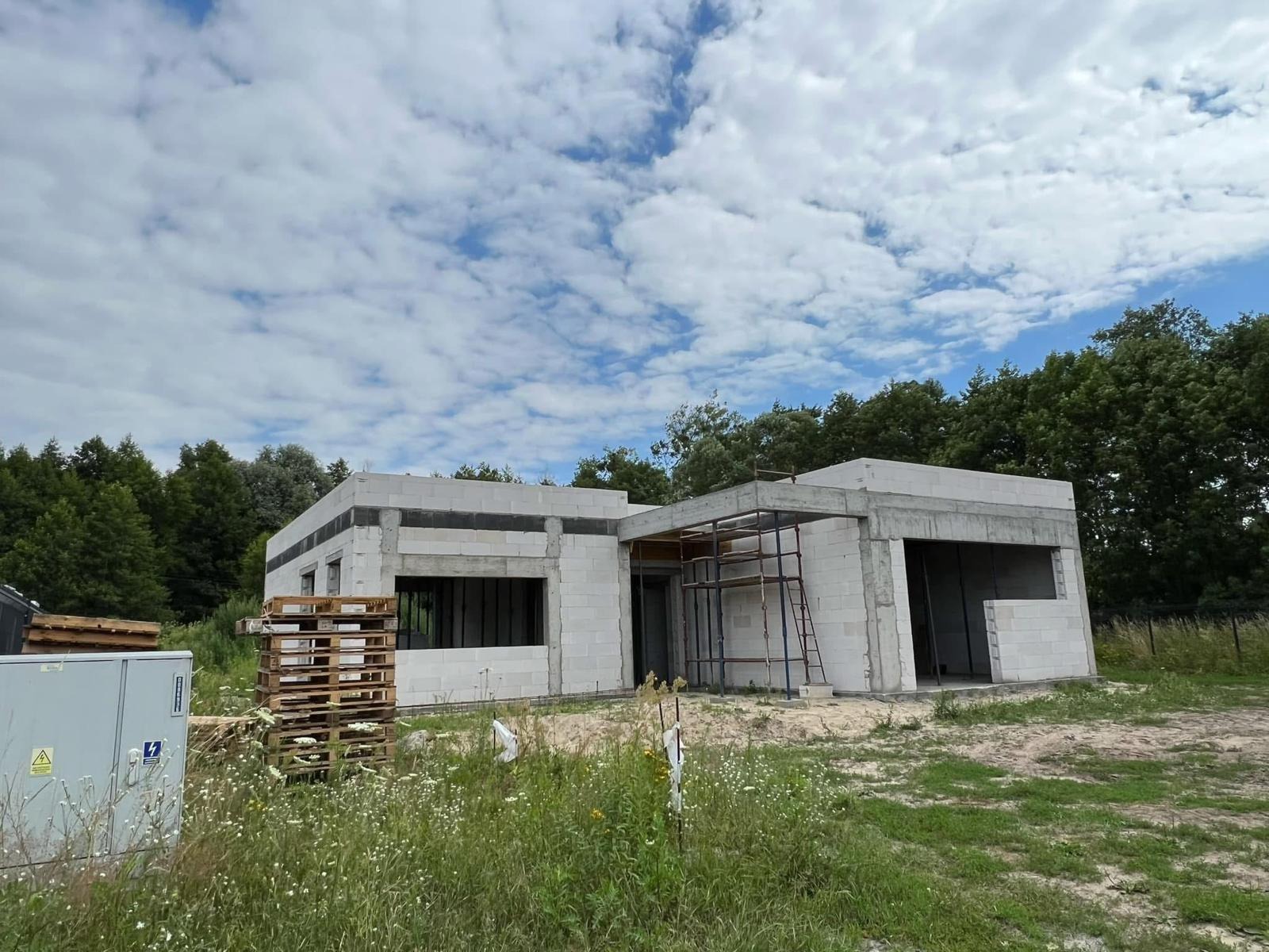 Budowa domu z bloczków betonu komórkowego w letni dzień. Widoczne rusztowania, palety z materiałem i bujna zieleń w tle. Stan surowy otwarty.