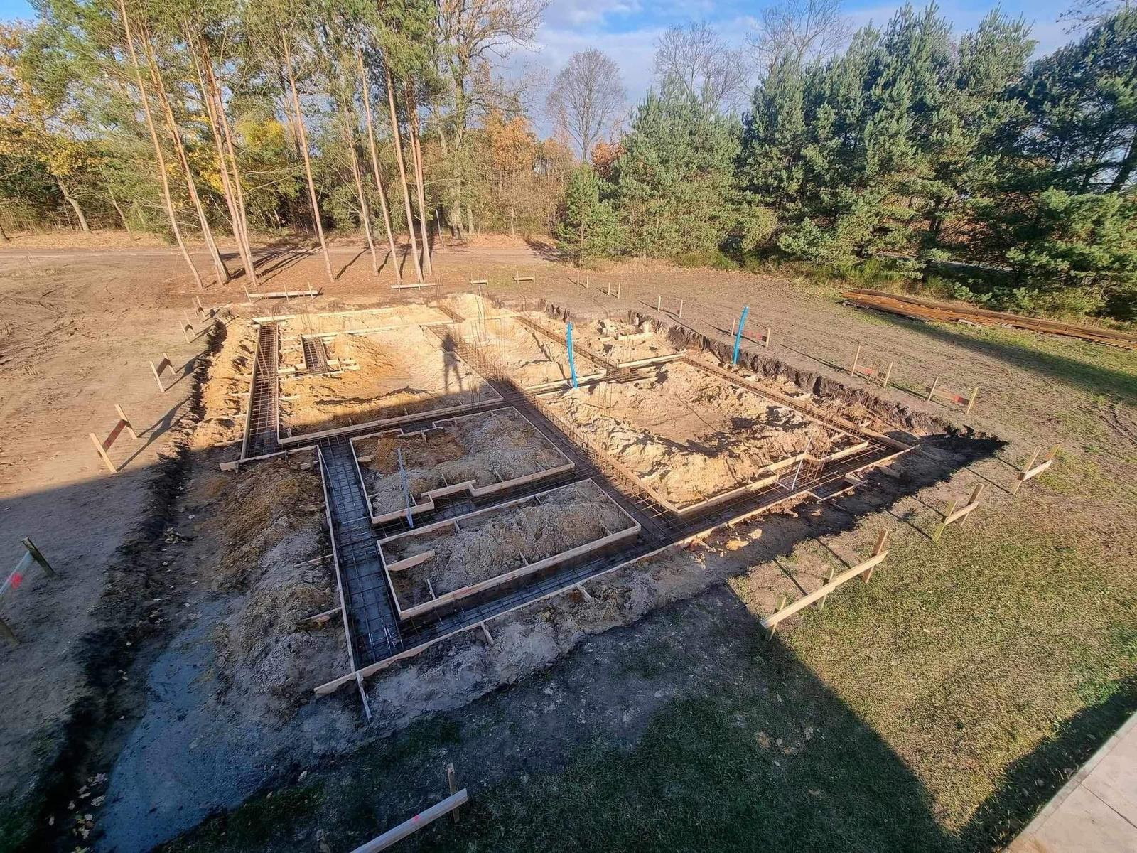 Zbrojenie fundamentów domu jednorodzinnego z drewnianym szalunkiem, widok z góry w słoneczny dzień. Wykopane fundamenty na działce z drzewami w tle.