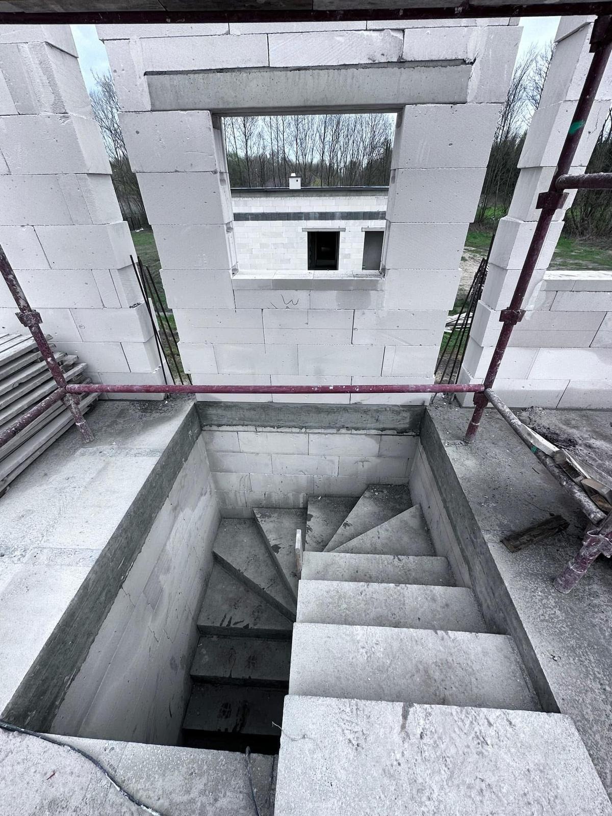 Wnętrze surowego domu w budowie: ściany z betonu komórkowego z otworem okiennym i betonowe schody prowadzące w dół. Widoczne rusztowania i elementy konstrukcyjne.