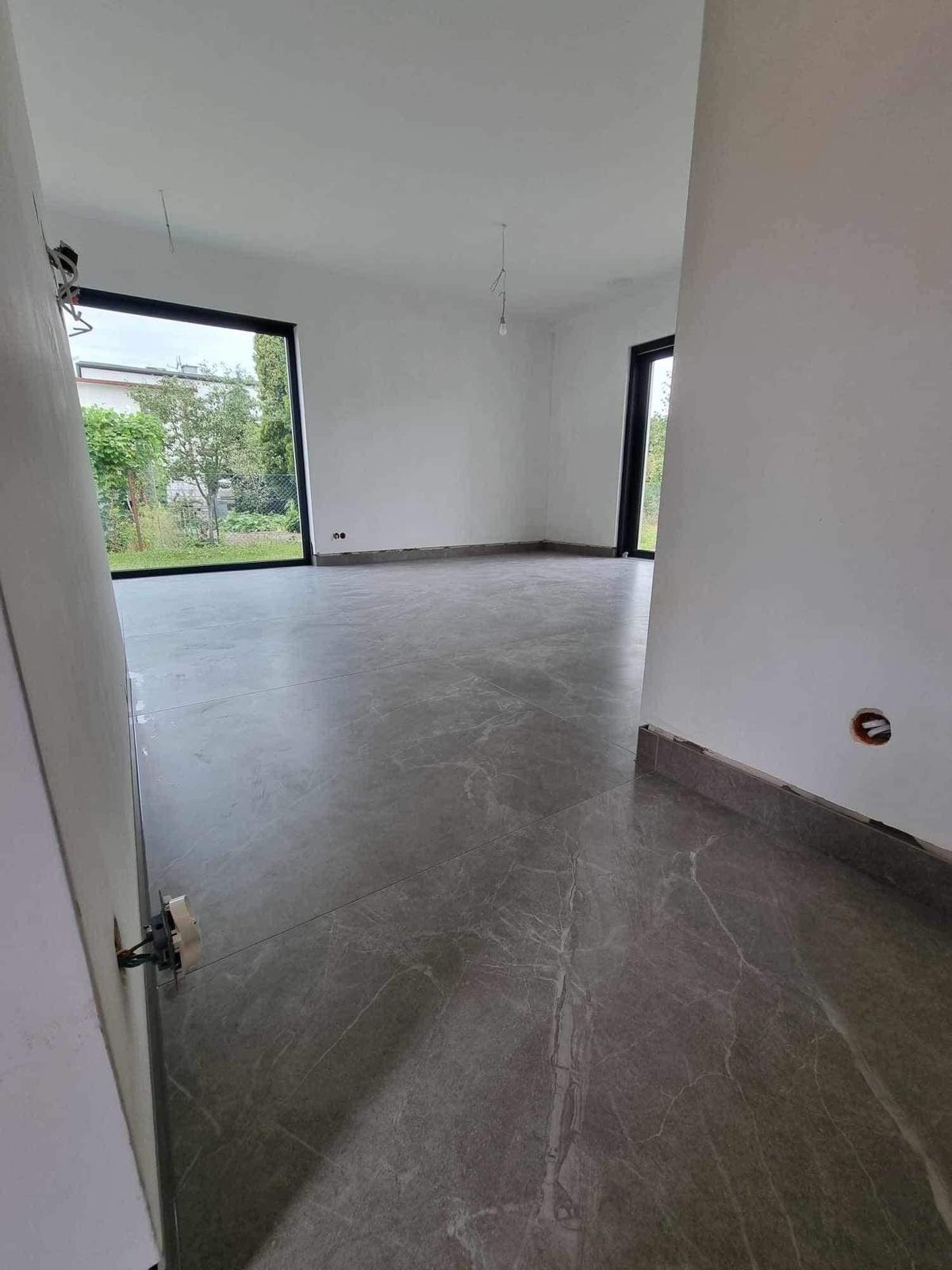 Wnętrze domu w stanie deweloperskim z szarą marmurową podłogą i dużymi oknami z widokiem na ogród. Widoczne niedokończone gniazdka elektryczne.
