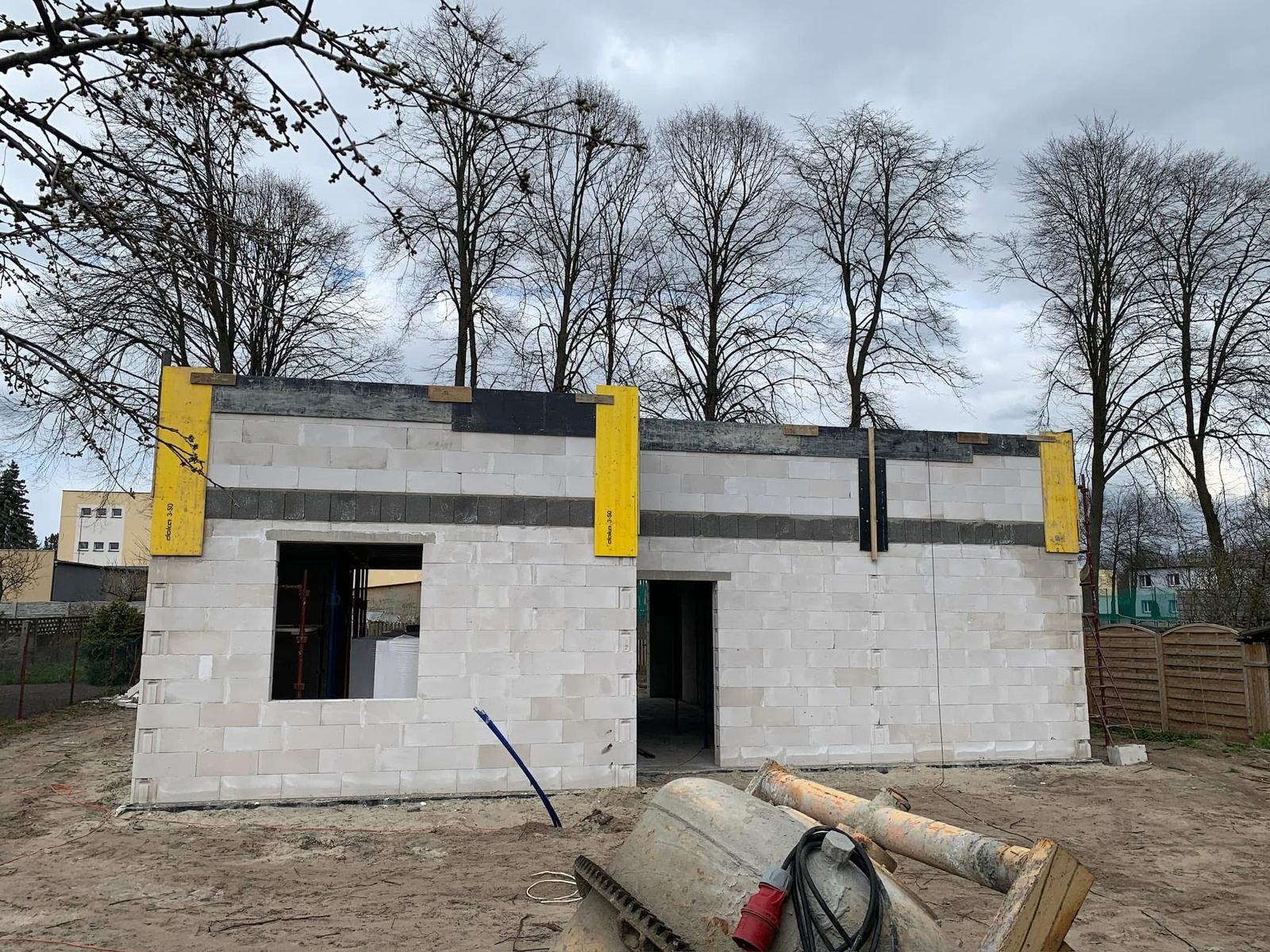 Wznoszenie murów domu z betonu komórkowego z widocznym oknem i otworem drzwiowym. Szalunki pionowe w kolorze żółtym. Na pierwszym planie betoniarka.