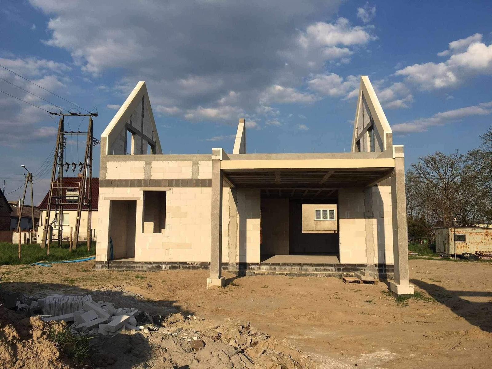 Surowy stan domu z betonu komórkowego w trakcie budowy, z charakterystycznymi szczytami i zadaszeniem werandy. W tle słupy energetyczne i zabudowa.