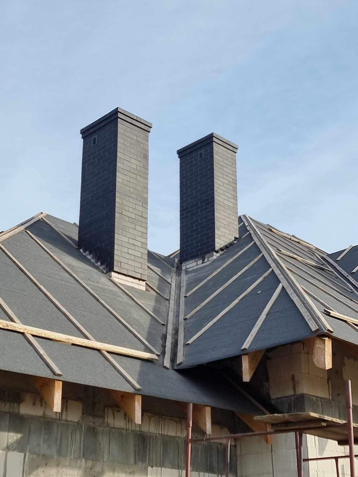 Ciemne kominy z cegły na dachu pokrytym papą, widoczne drewniane łaty i fragment ściany w trakcie budowy. Ujęcie z dołu, błękitne niebo w tle.