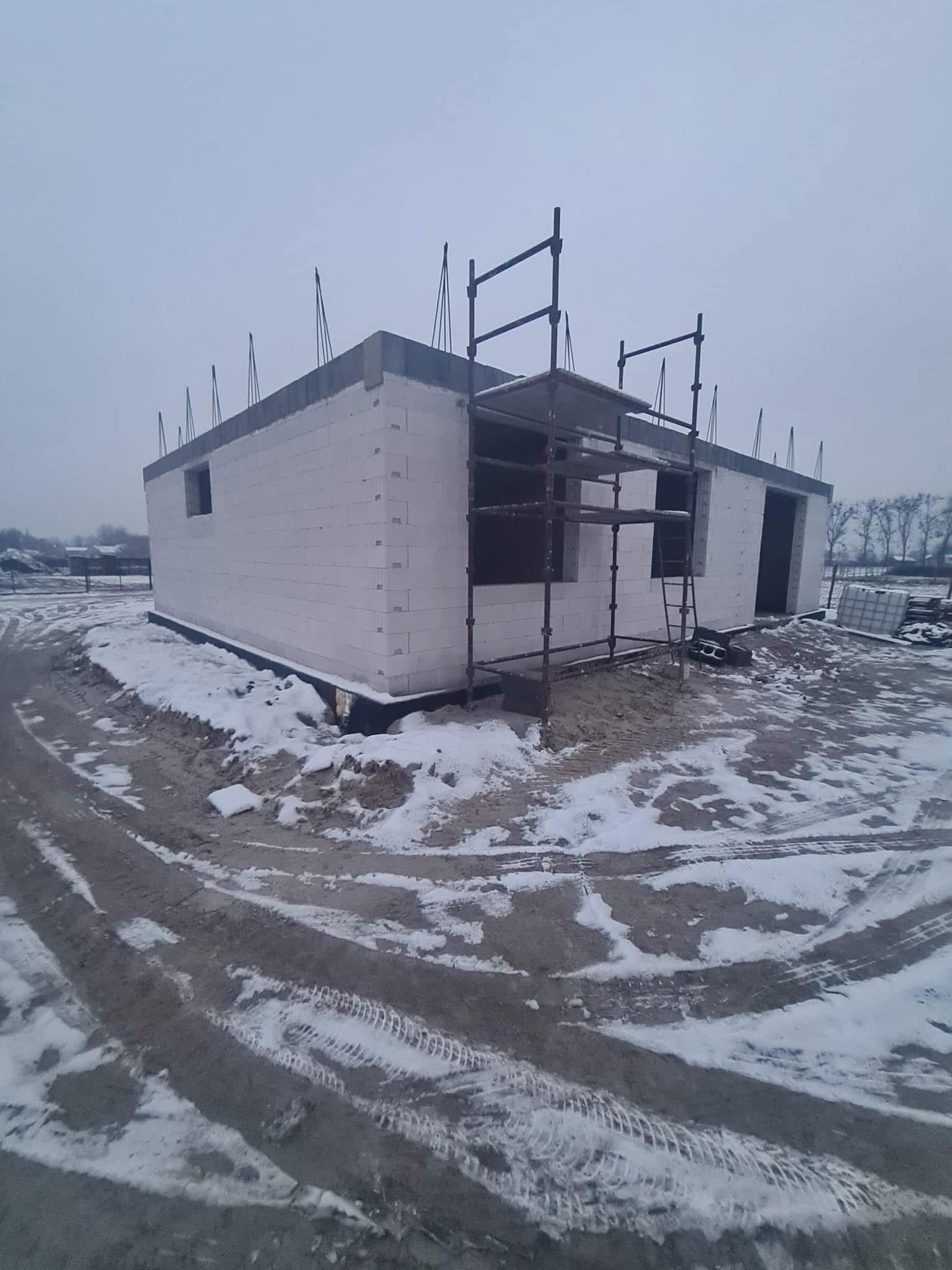 Surowy stan domu z białego pustaka, z widocznymi oknami i drzwiami. Na zewnątrz rusztowanie i ślady opon na śniegu, pochmurna pogoda.