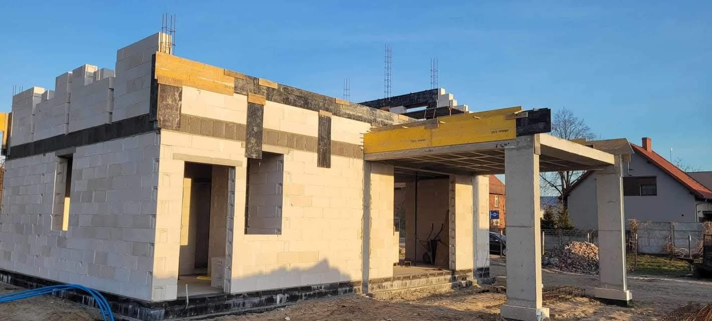 Niedokończony dom z betonu komórkowego z widoczną konstrukcją nośną, deskowaniem i zbrojeniami. W tle niebieskie niebo i zabudowa jednorodzinna.