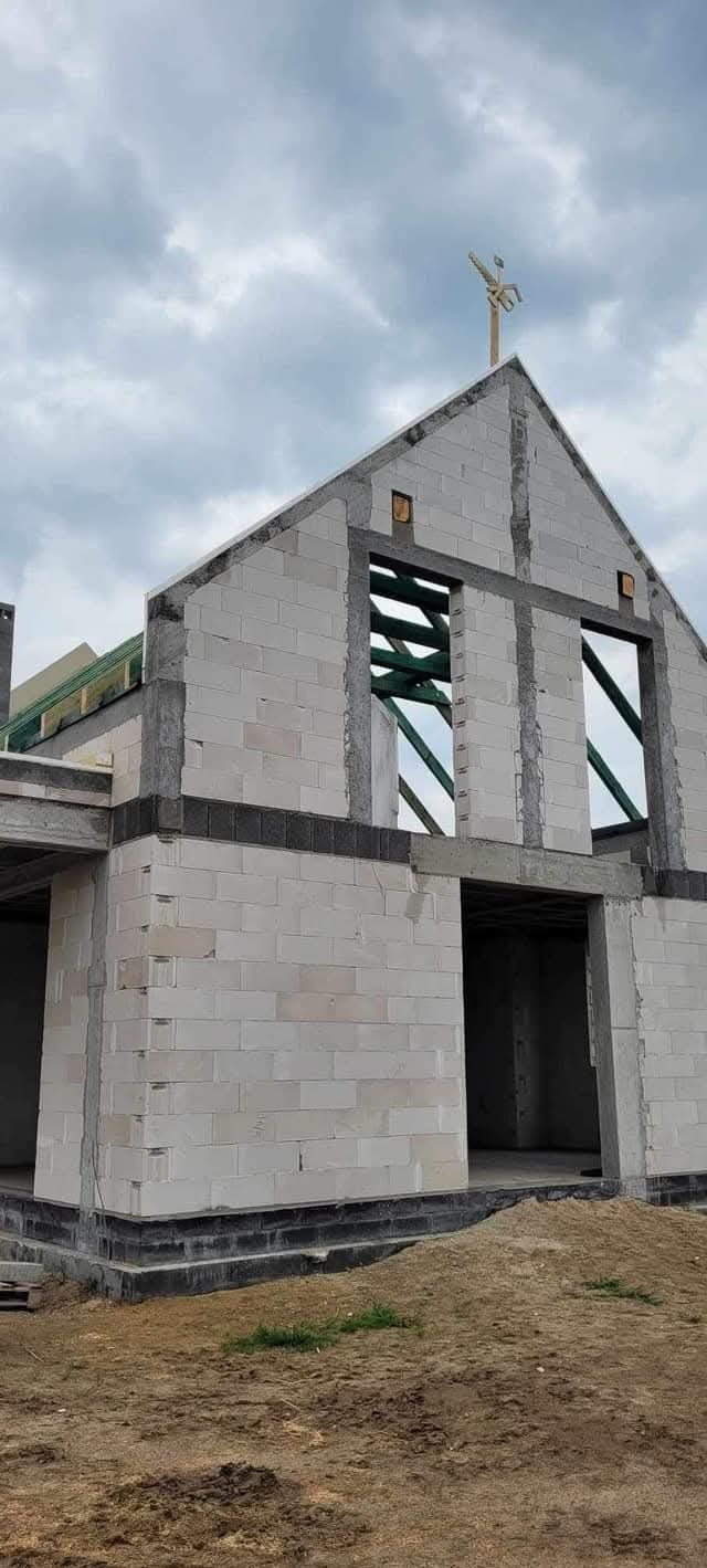 Surowy stan domu z betonu komórkowego, widoczne otwory okienne i drzwiowe, konstrukcja dachu w trakcie budowy, krzyż na dachu, pochmurne niebo w tle.