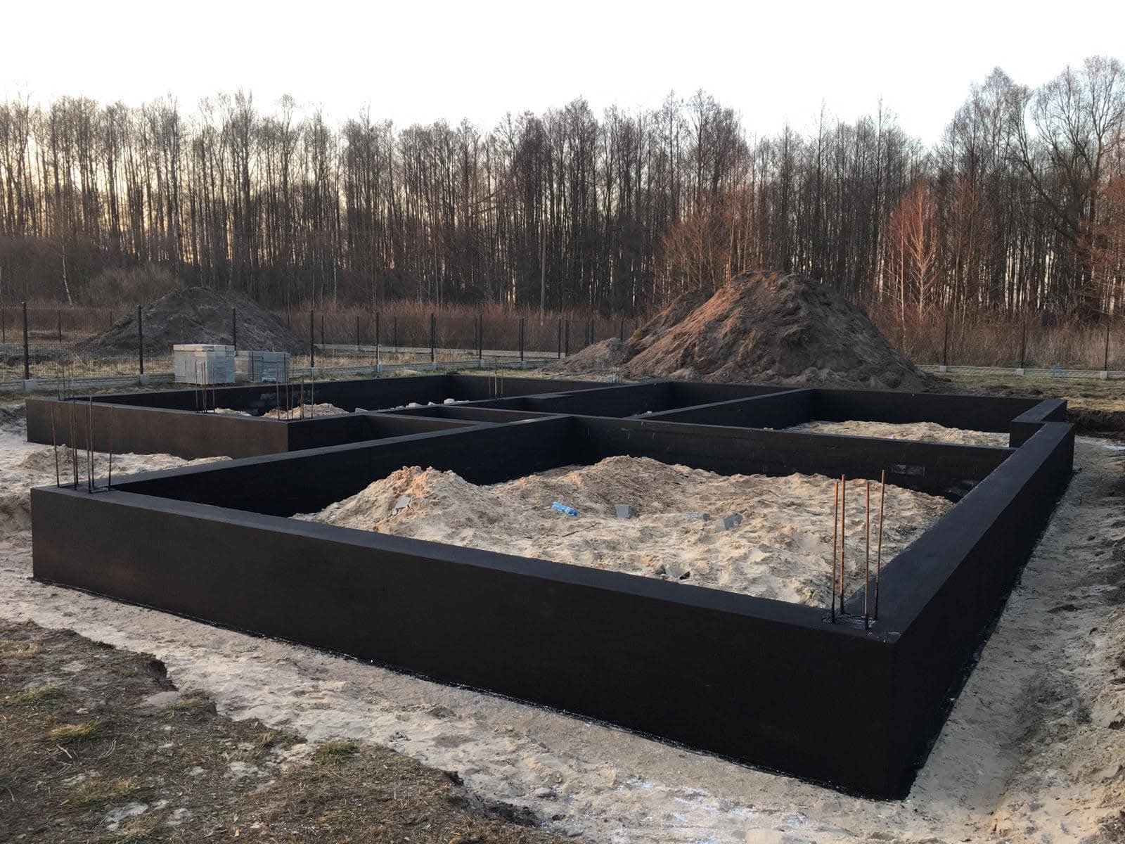 Zalane fundamenty domu jednorodzinnego, widoczne zbrojenia, w tle hałda piachu i las. Czarna izolacja przeciwwilgociowa na betonie.