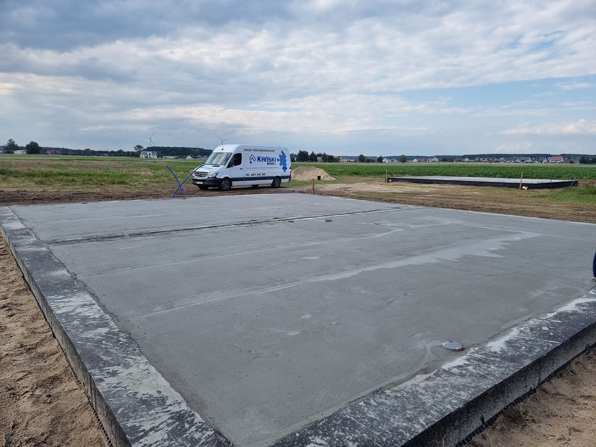 Wylana płyta fundamentowa na budowie domu jednorodzinnego, z widocznym firmowym vanem w tle, na tle zielonego pola pod pochmurnym niebem.