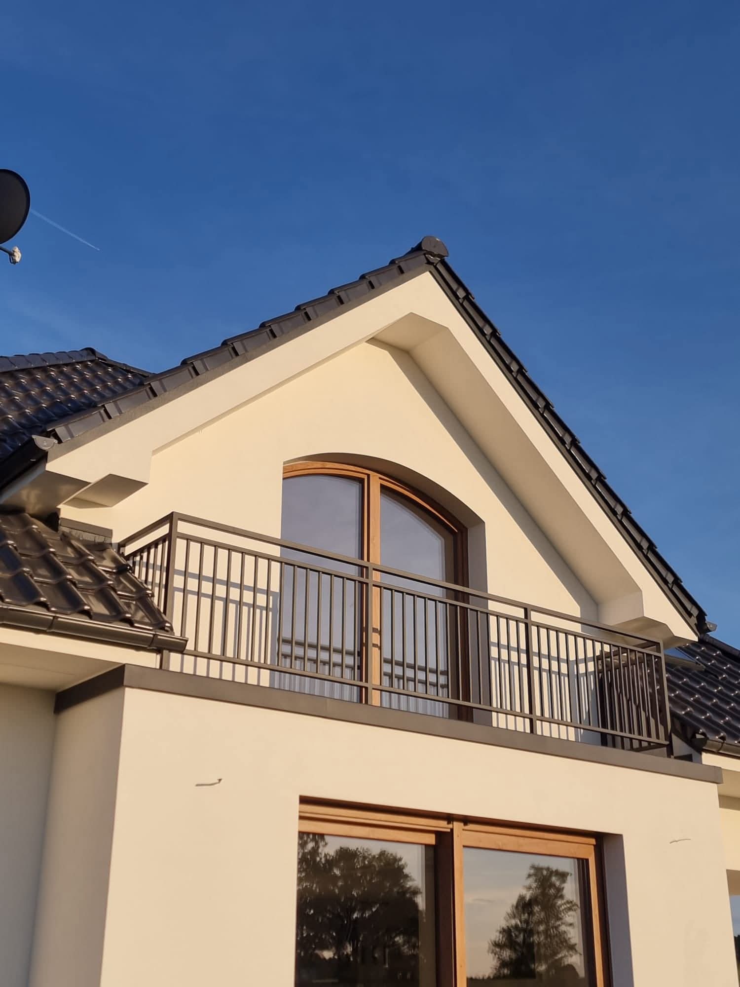 Nowoczesny dom z balkonem, jasna elewacja, grafitowa balustrada i dachówka. Okno balkonowe z łukiem. Błękitne niebo w tle, widoczny ślad po samolocie.