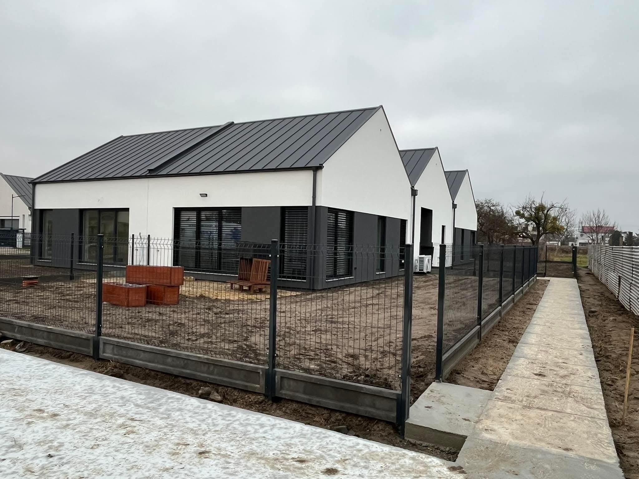 Nowoczesne domy jednorodzinne z szarymi dachami, widok z zewnątrz, ogrodzenie panelowe z podmurówką, elementy małej architektury ogrodowej, pochmurne niebo.