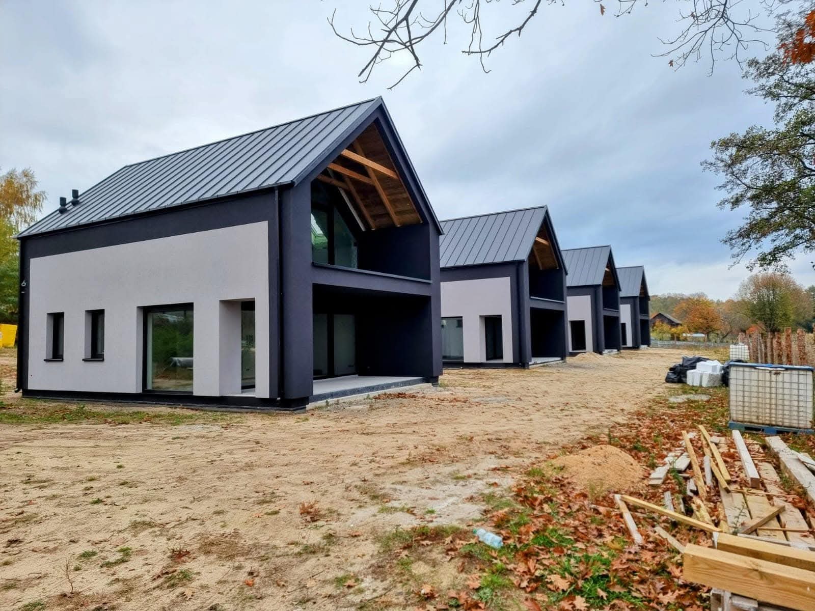 Szereg identycznych, nowoczesnych domów z dwuspadowymi dachami i ciemnymi elewacjami, widok z zewnątrz, jesienne liście na ziemi, pochmurne niebo.