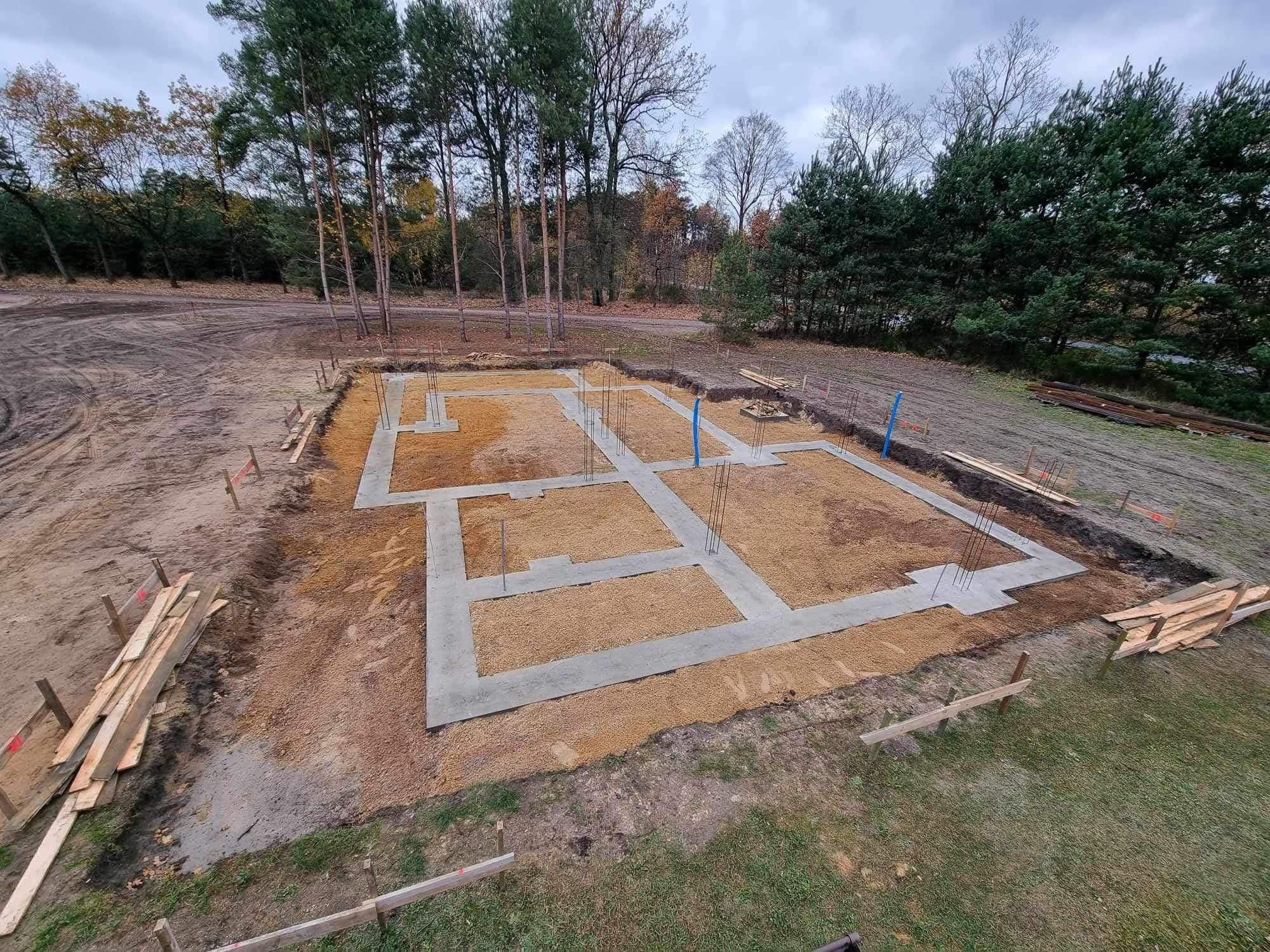 Zalane betonem fundamenty domu jednorodzinnego z wystającymi prętami zbrojeniowymi. Widok z góry na wykopane i przygotowane podłoże z deskami szalunkowymi.