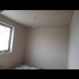 Konrad Kocemba - Pokój po nałożeniu gładzi. Ściany i sufit są świeżo wygładzone, widoczne gruz i resztki na podłodze. Okno z częściowo usuniętą stolarką, surowa żarówka.