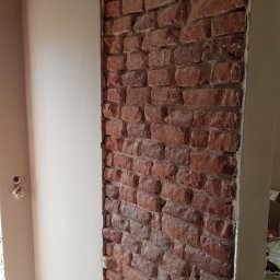 Konrad Kocemba - Częściowo otynkowana ściana z odsłoniętą czerwoną cegłą, widoczne gniazdka elektryczne. Prace wykończeniowe wnętrza.