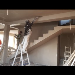 Konrad Kocemba - Dwóch mężczyzn na budowie podczas nakładania gładzi na ścianę i sufit w jasnym pomieszczeniu z widocznymi schodami i drabinami. Praca w toku.