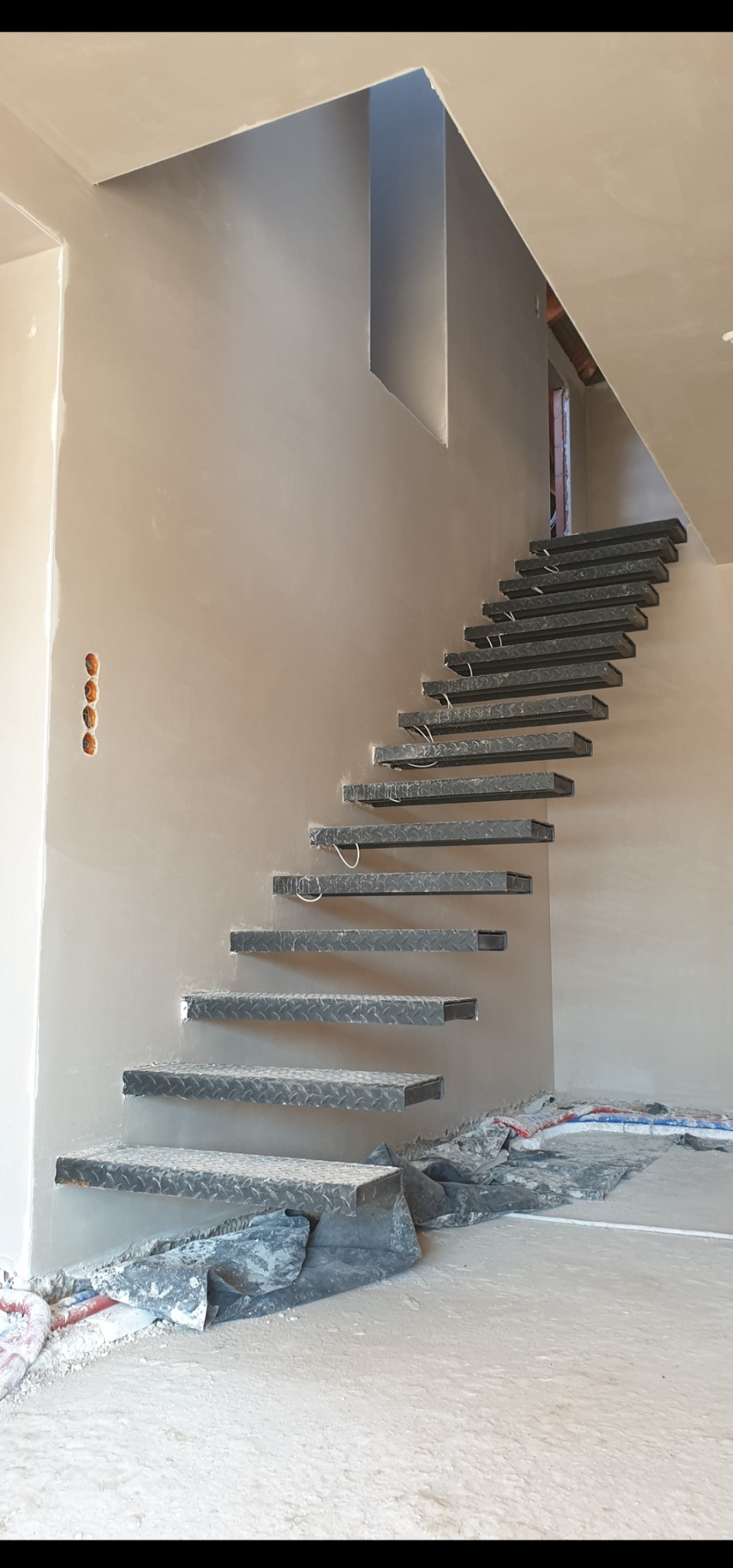 Surowe, metalowe schody wspornikowe w jasnym wnętrzu w trakcie wykańczania. Ściany pokryte świeżą, jasną gładzią, widoczne otwory pod gniazdka elektryczne.