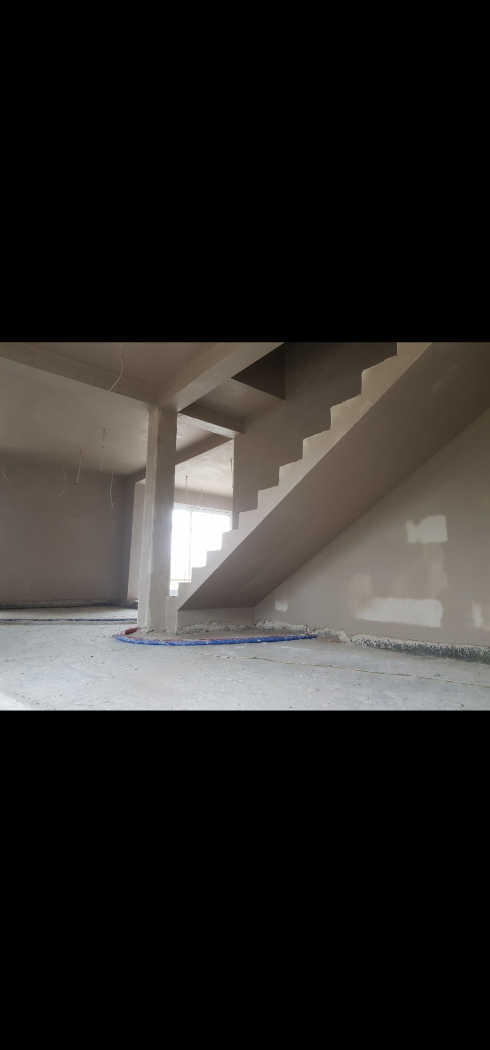 Wnętrze w stanie surowym: ściany z gładzią, schody, widoczne instalacje podłogowe. Duże okno w tle. Jasne, naturalne światło.