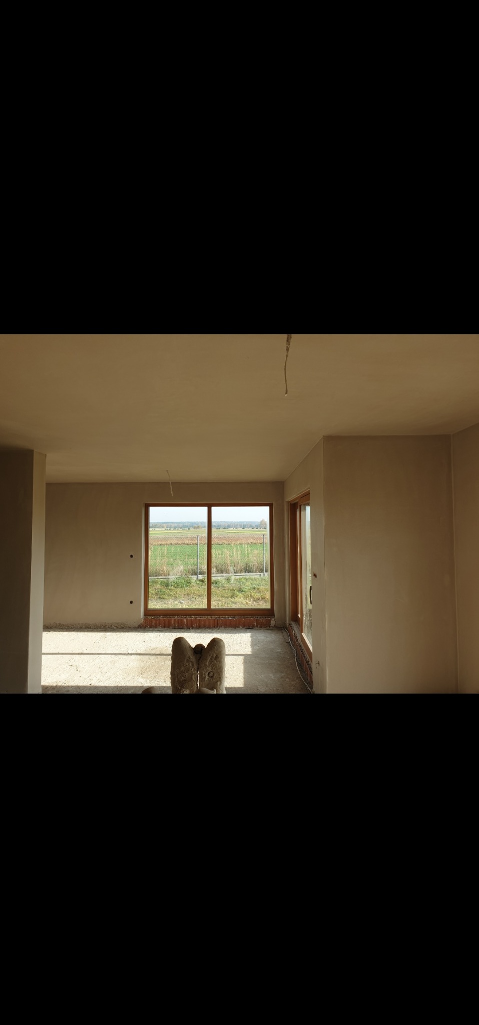 Wnętrze po tynkowaniu maszynowym, widok na okno z ceglanym parapetem. Ściany gładkie, jasne, przygotowane do dalszych prac wykończeniowych. W tle widok na pola.