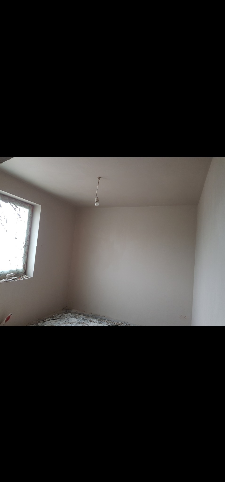 Pokój po nałożeniu gładzi. Ściany i sufit są świeżo wygładzone, widoczne gruz i resztki na podłodze. Okno z częściowo usuniętą stolarką, surowa żarówka.