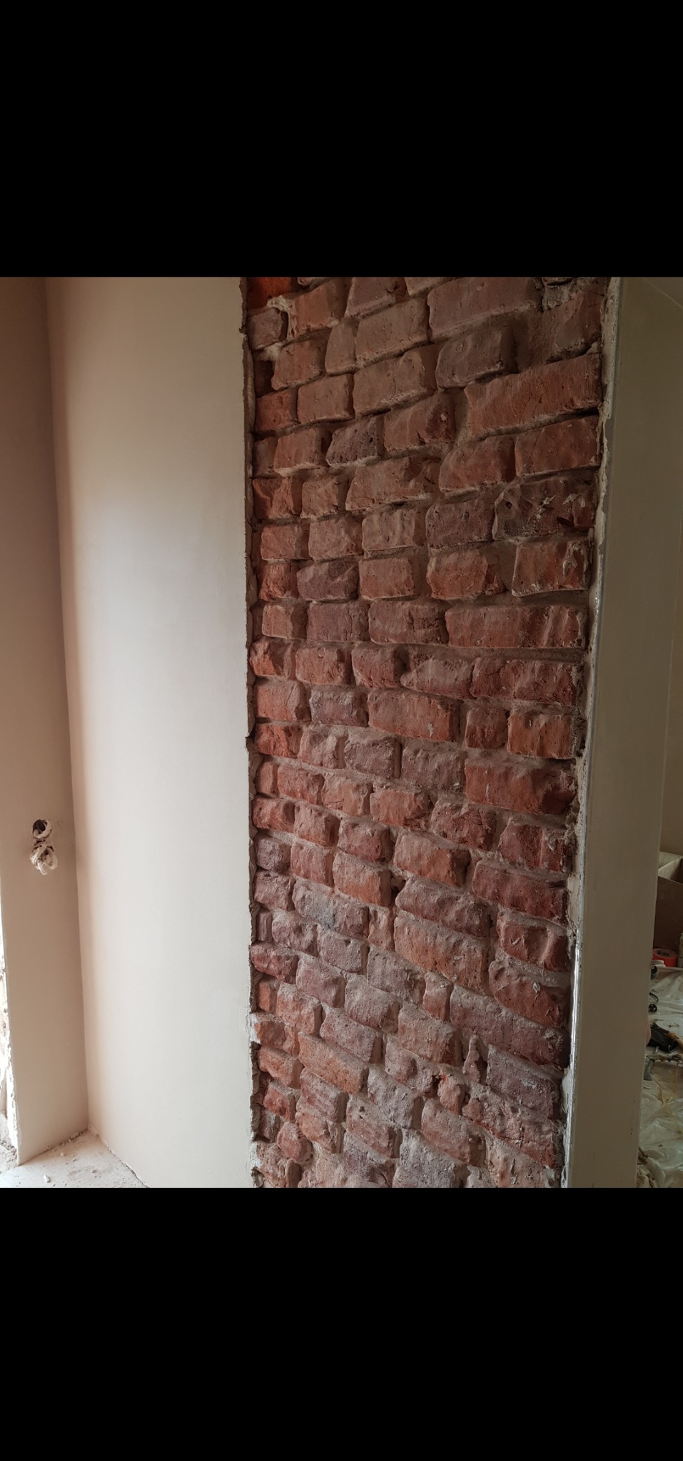 Częściowo otynkowana ściana z odsłoniętą czerwoną cegłą, widoczne gniazdka elektryczne. Prace wykończeniowe wnętrza.