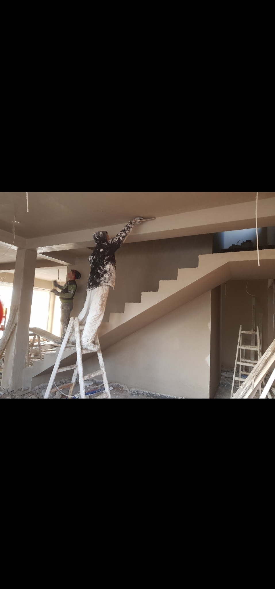 Dwóch mężczyzn na budowie podczas nakładania gładzi na ścianę i sufit w jasnym pomieszczeniu z widocznymi schodami i drabinami. Praca w toku.