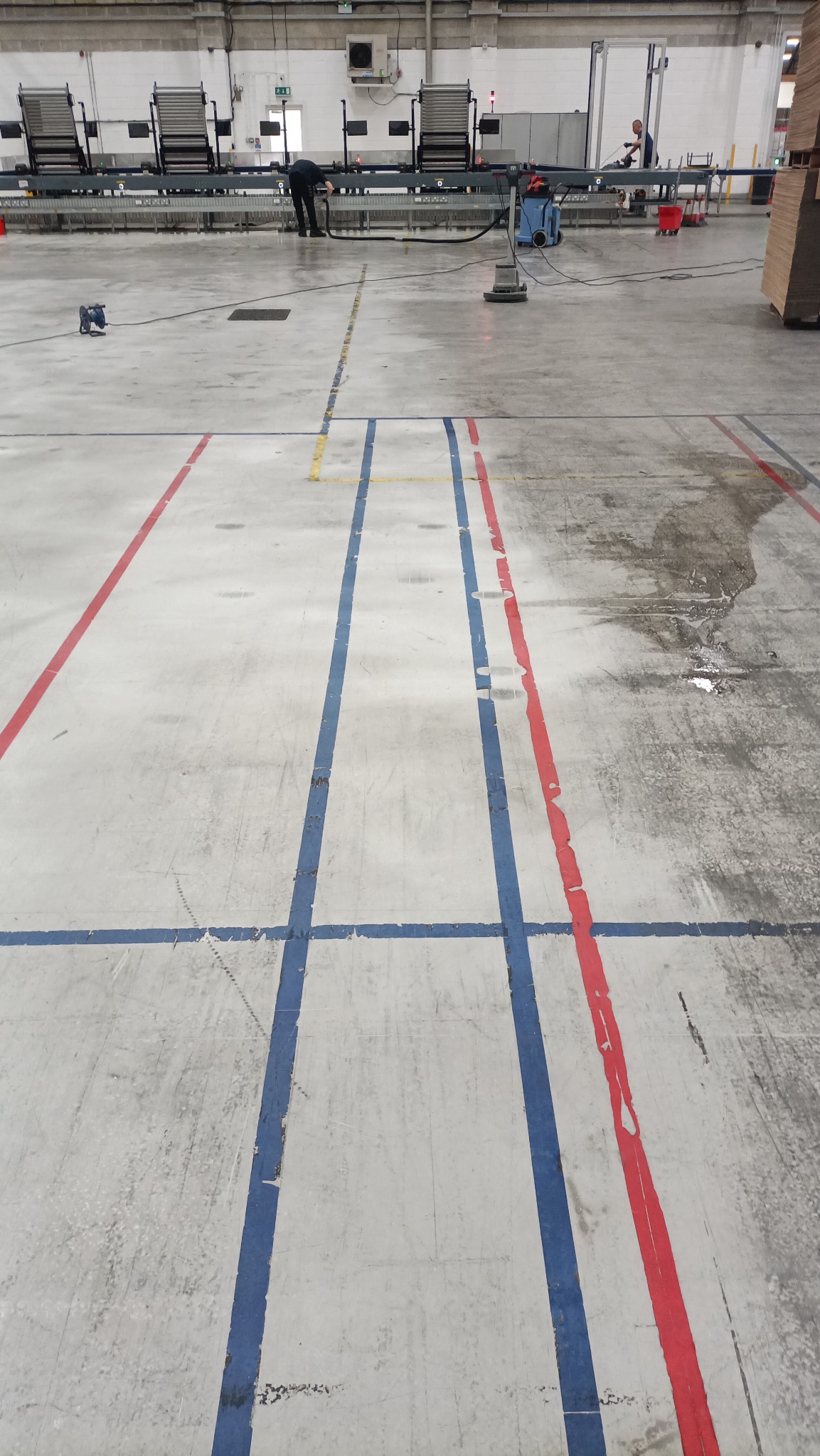 Czyszczenie posadzki w hali przemysłowej z liniami. Widoczne maszyny czyszczące i pracownicy w tle, podkreślające skalę przestrzeni i etap prac porządkowych.