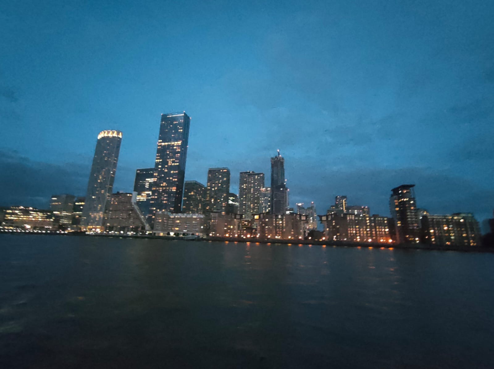 Nocny widok na panoramę miasta z rzeki, z licznymi oświetlonymi wieżowcami i budynkami mieszkalnymi, odbijającymi się w wodzie pod ciemnym, pochmurnym niebem.