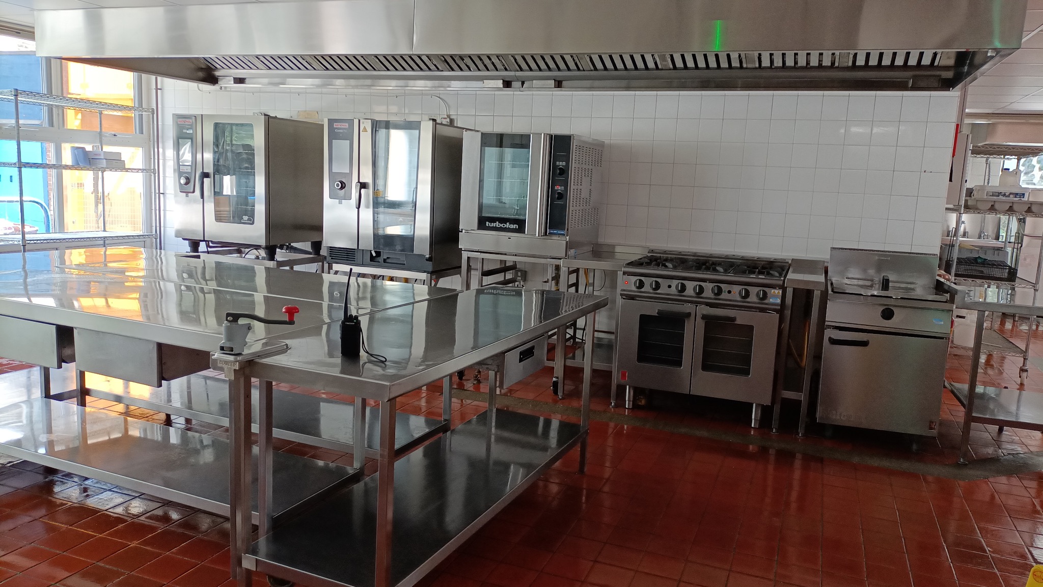 Profesjonalne wyposażenie kuchni: stalowe stoły, piece konwekcyjne i kuchenka gazowa pod okapem w lokalu gastronomicznym. Widoczne płytki na ścianach i podłodze.