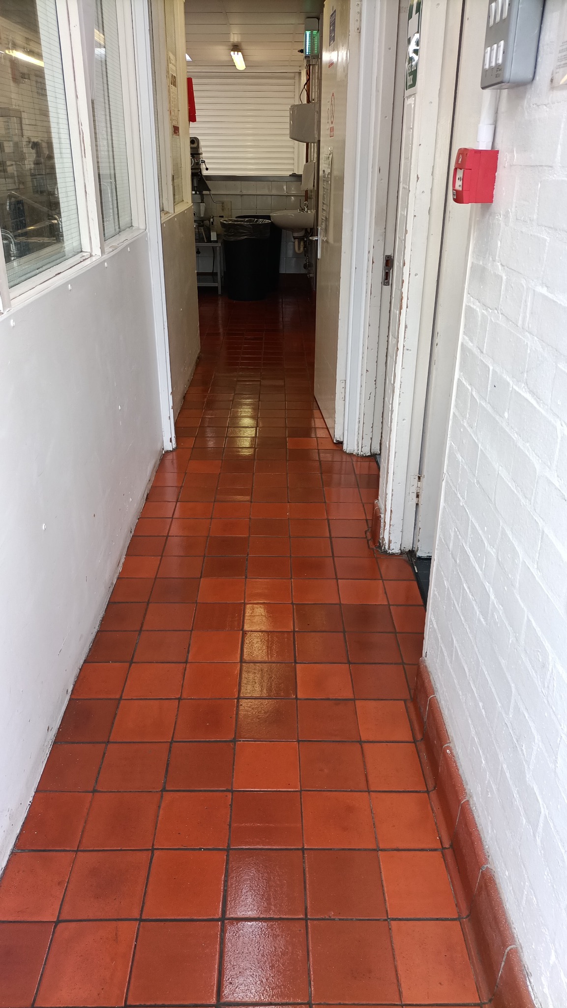 Błyszcząca, terakotowa podłoga w korytarzu z białymi ścianami i widocznym fragmentem zaplecza w tle. Widoczne ślady po umyciu podłogi.
