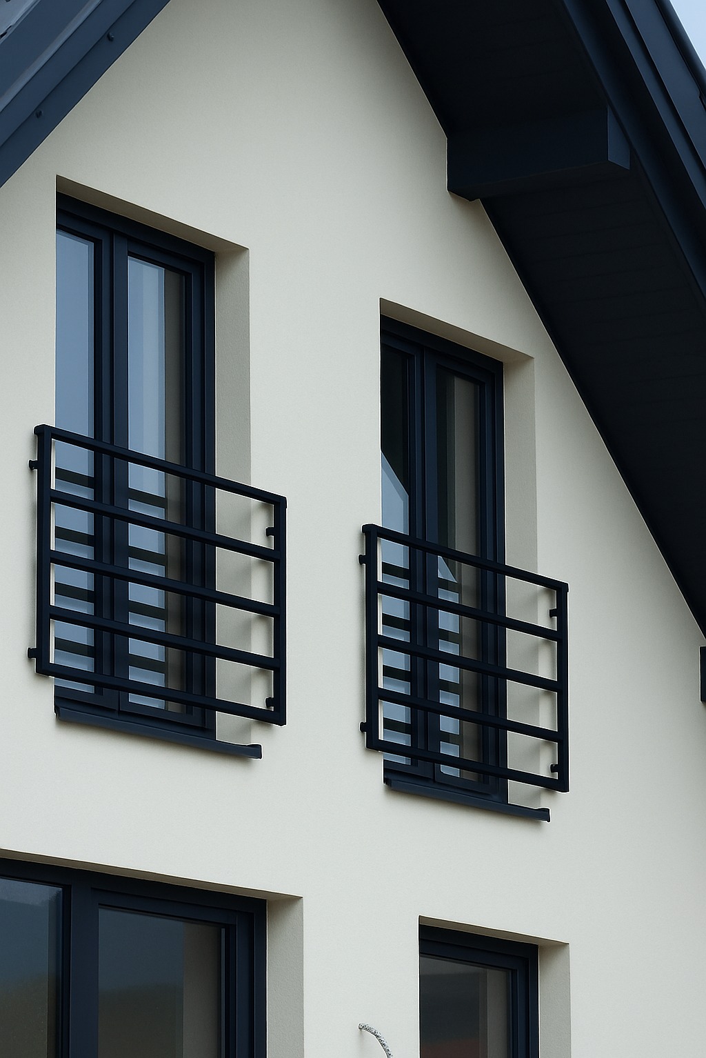 Elewacja domu z dwoma oknami zabezpieczonymi czarnymi, metalowymi konstrukcjami, pełniącymi funkcję balustrad.
