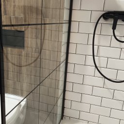 Firma sprzątająca - Szczegółowy widok kabiny prysznicowej z czarną armaturą i półką z kosmetykami na tle białych, prostokątnych płytek. Widoczne uszczelnienia i silikon w narożnikach.