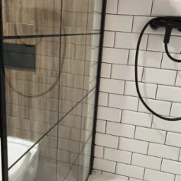 Szczegółowy widok kabiny prysznicowej z czarną armaturą i półką z kosmetykami na tle białych, prostokątnych płytek. Widoczne uszczelnienia i silikon w narożnikach.