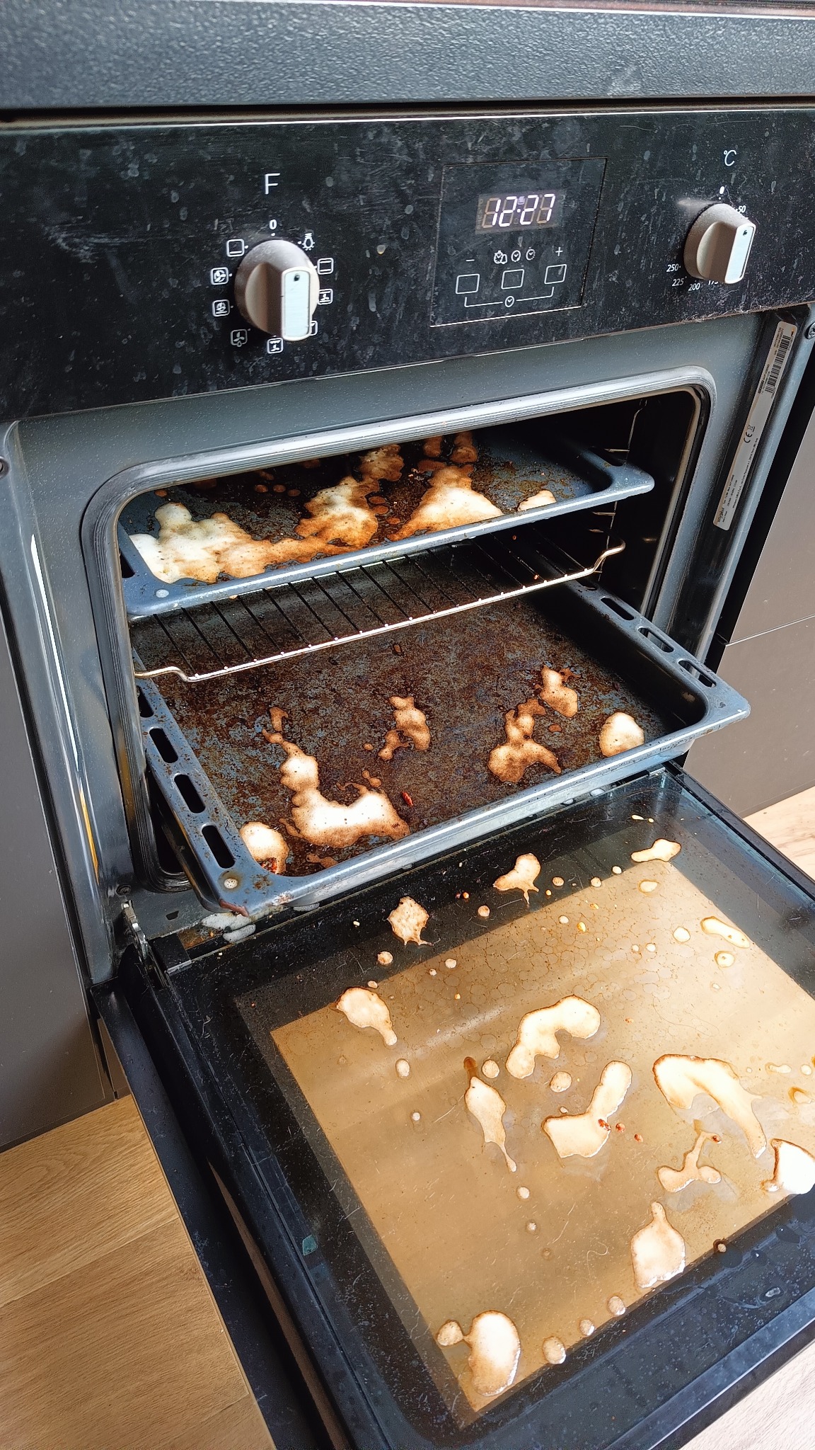 Zabrudzony piekarnik z resztkami jedzenia na blachach i szybie, wymagający gruntownego czyszczenia. Widoczne przypalone plamy i osady.