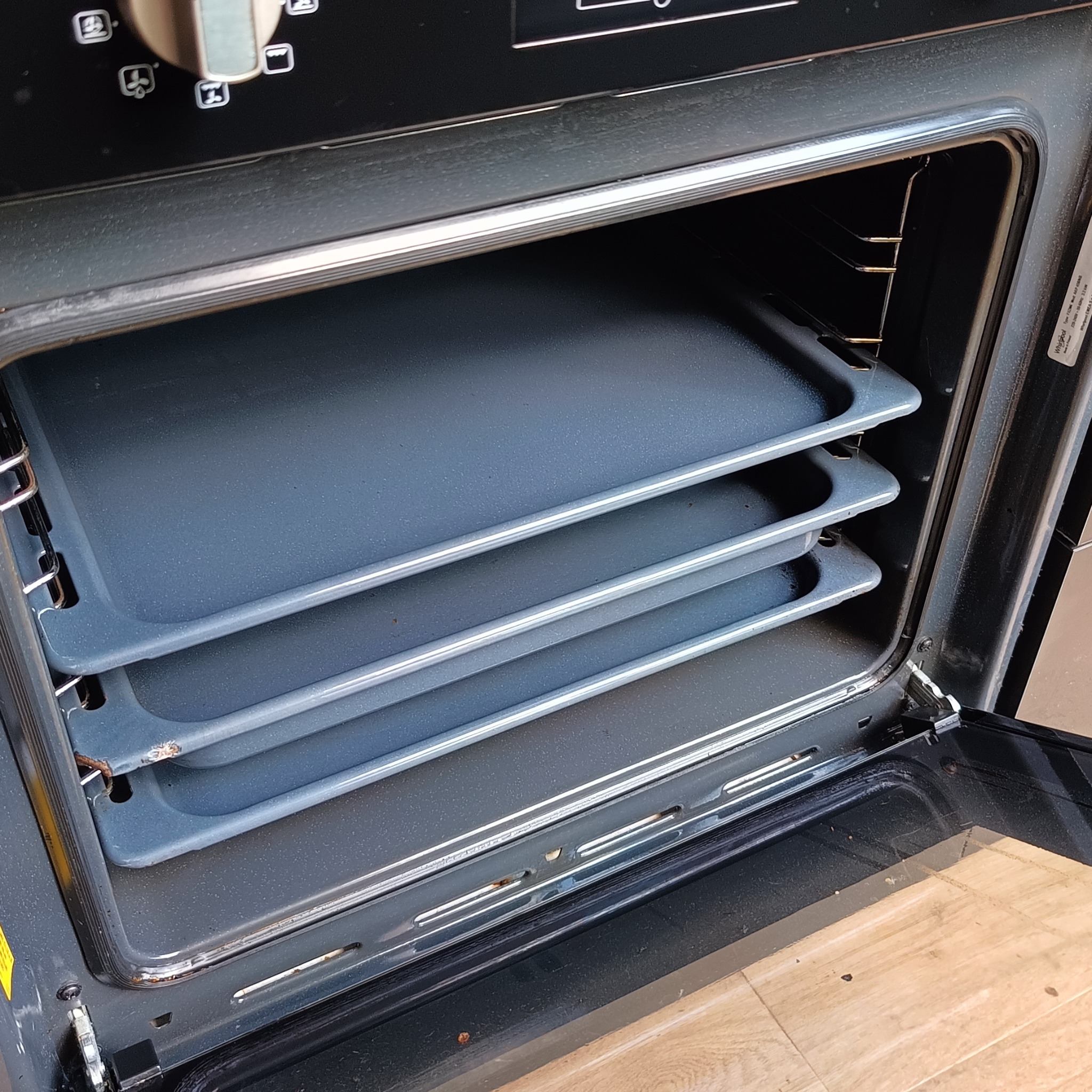 Otwarty piekarnik z trzema blachami w środku. Widoczne ślady użytkowania na wewnętrznych ściankach i blachach. Oświetlenie wnętrza piekarnika.