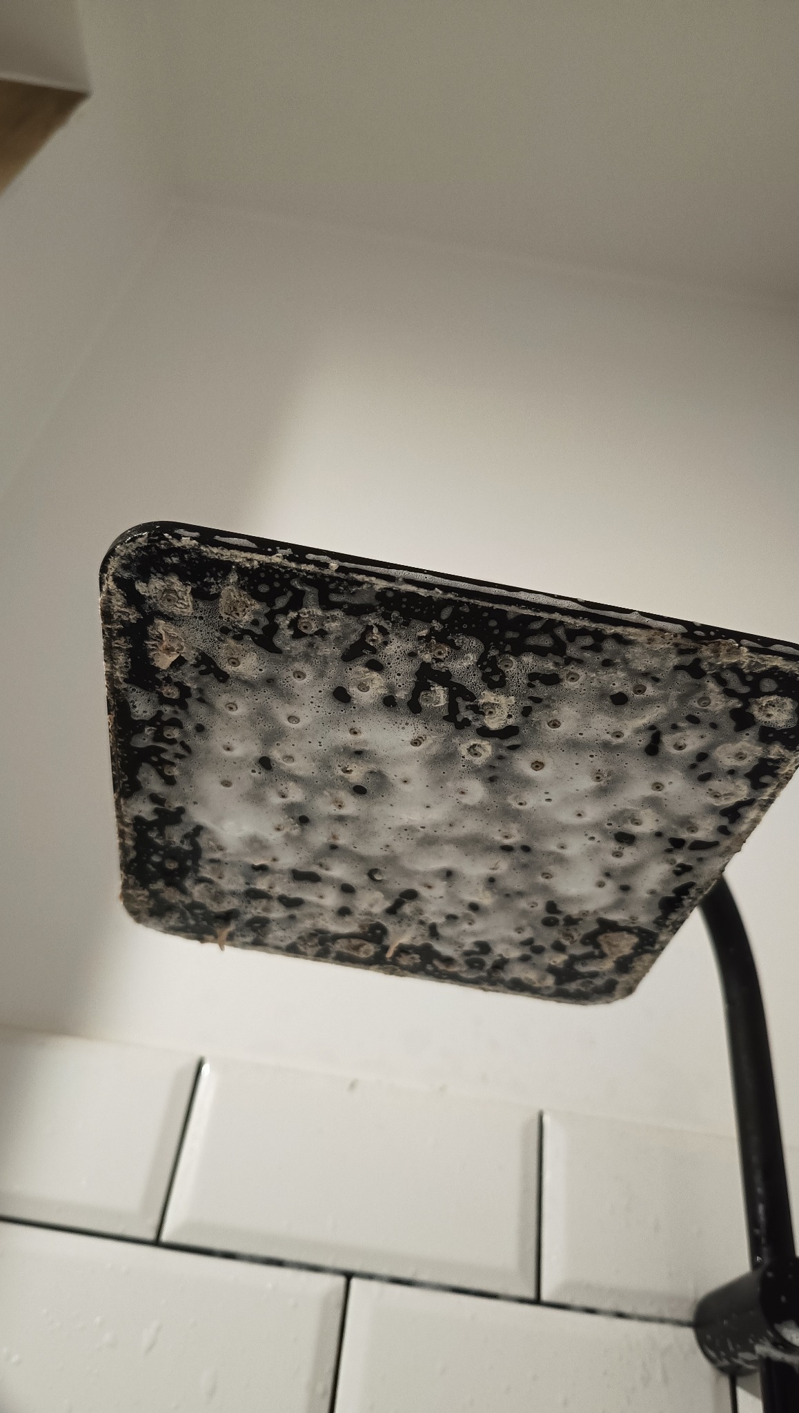 Zanieczyszczona, czarna głowica prysznicowa pokryta pianą i osadem, widoczna na tle białych płytek w łazience. Silne zabrudzenia wymagają dokładnego czyszczenia.