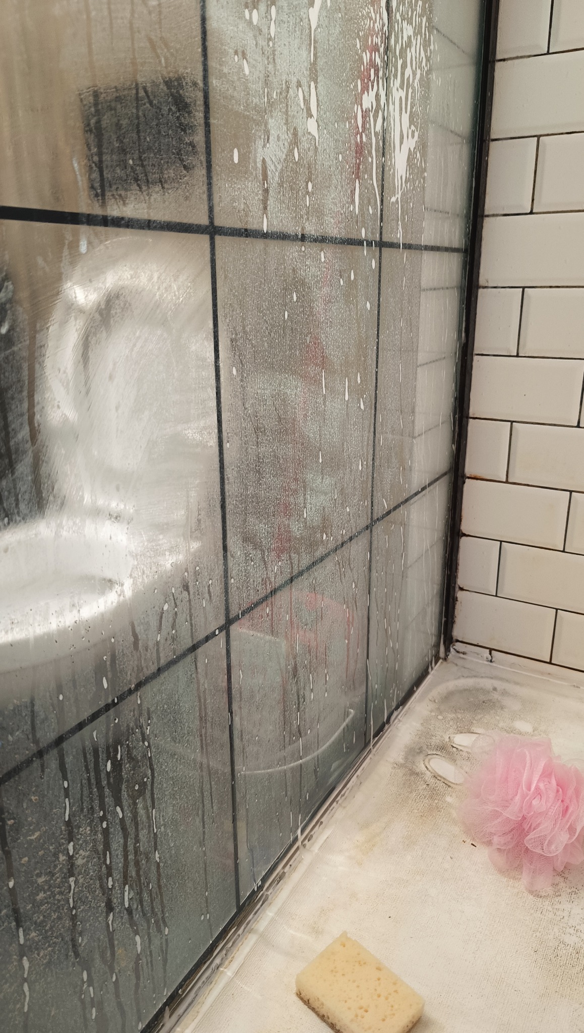 Piana i zacieki na szklanej kabinie prysznicowej z czarnymi ramami, gąbka i myjka na brodziku. Widoczne białe płytki na ścianie.