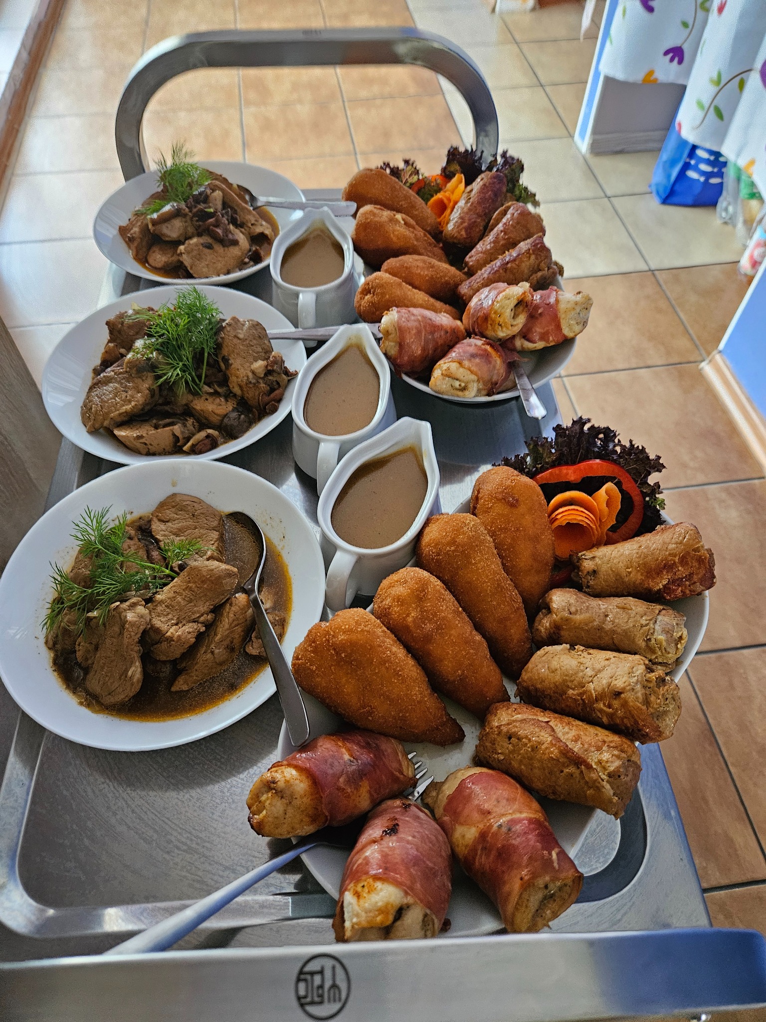 Metalowy wózek cateringowy z gotowymi daniami: mięso w sosie, krokiety, roladki w szynce, sosjerkami. Dekoracje z warzyw, świeży koperek. Idealne na imprezę firmową.