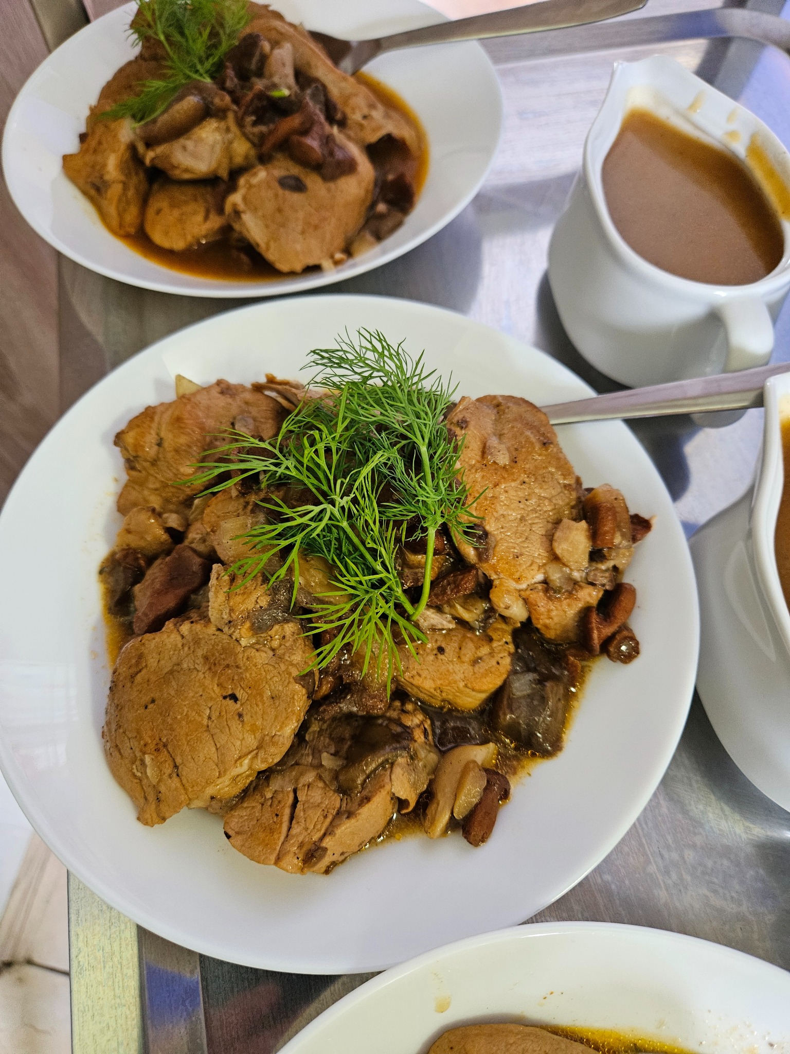 Dania mięsne z sosem i koperkiem na białych talerzach, widoczne sosjerki. Potrawy cateringowe gotowe do serwowania na przyjęciu.