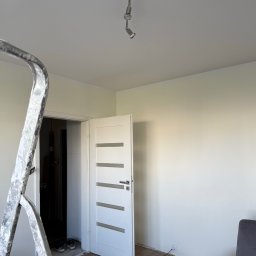 Usługi Remontowo Wykończeniowe Filip Hacz - Wnętrze po malowaniu ścian na jasny odcień, widoczna drabina malarska, białe drzwi z przeszkleniami i fragment sofy. Parkiet na podłodze.