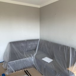 Usługi Remontowo Wykończeniowe Filip Hacz - Pokój przygotowany do malowania: narożnik przykryty folią ochronną, widoczne drabina i wałki malarskie. Ściany w odcieniach szarości, listwa sufitowa biała.