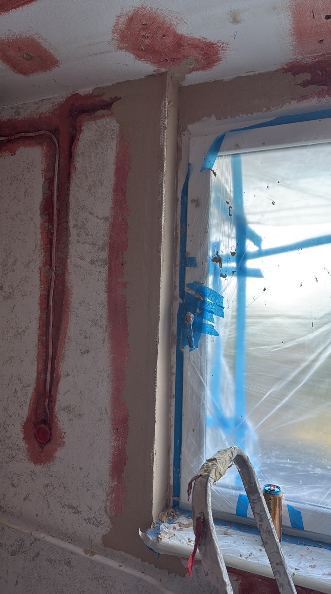 Ściana po szpachlowaniu z widocznymi śladami po instalacji elektrycznej, okno zabezpieczone folią i drabina. Etap przygotowania do malowania.