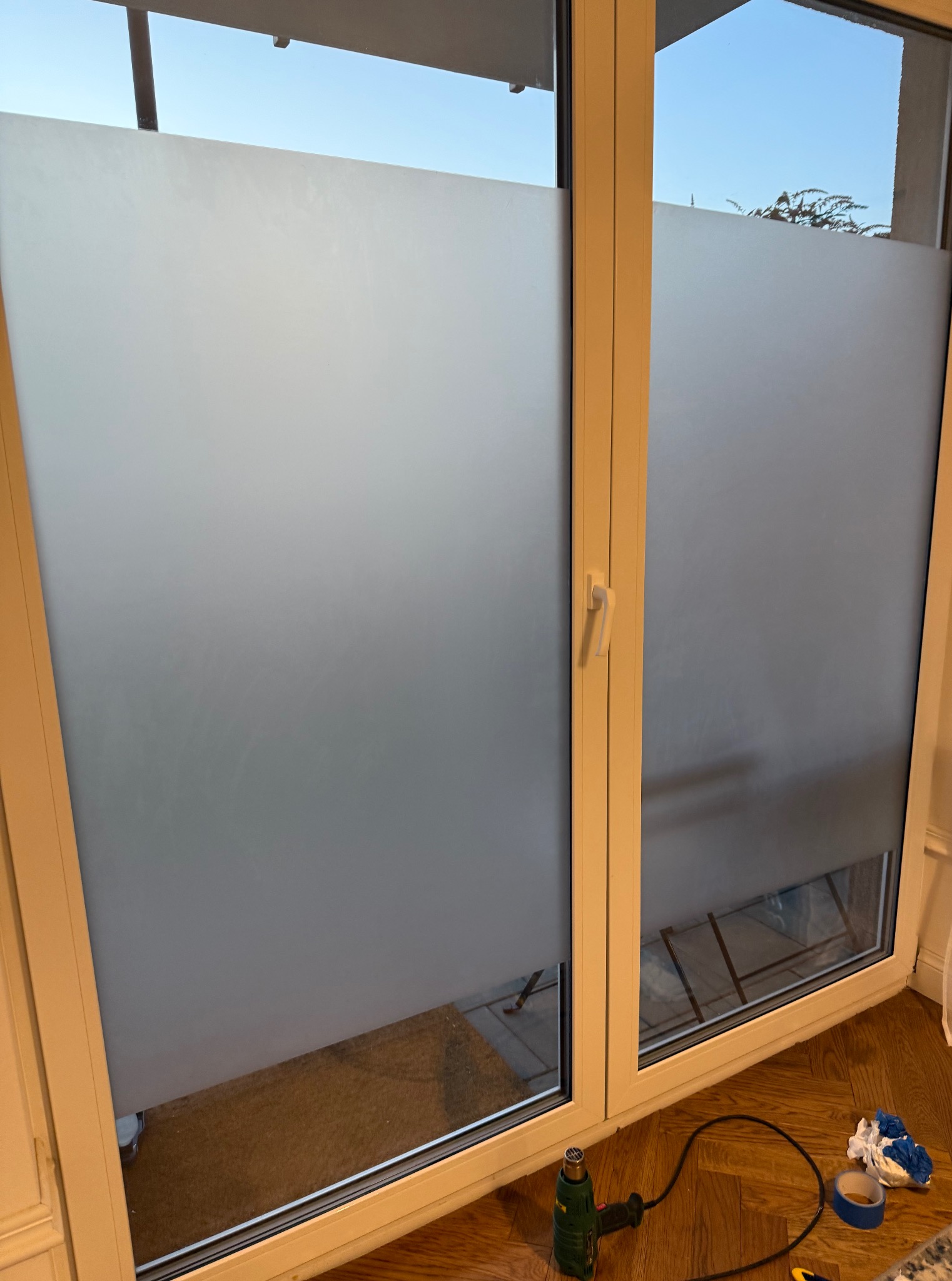 Okno z naklejoną folią matującą, widoczne narzędzia do aplikacji folii. Częściowe zaciemnienie okna dla prywatności lub dekoracji.