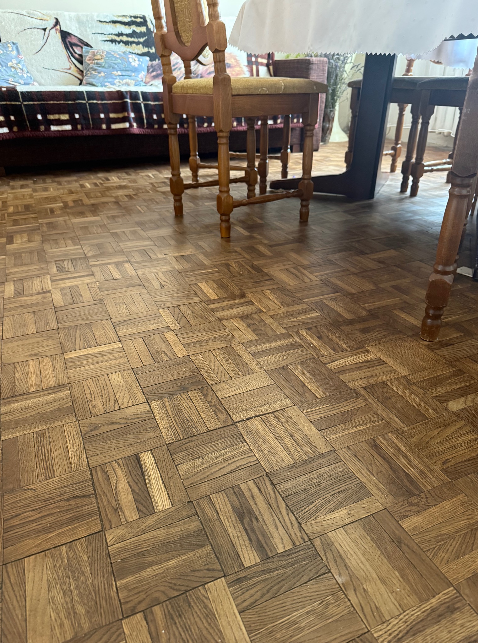 Parkiet w kwadraty z drewna dębowego, ułożony w jodełkę, wypełnia kadr. W tle widoczny fragment sofy i krzesła, co sugeruje wnętrze mieszkalne.